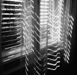 Black and white image of a window with suns light streaming through blind slats 