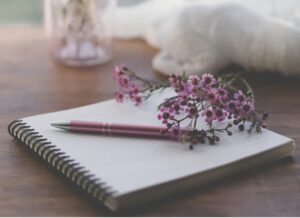 Image shows an open notebook with a pen resting on it, and a sprig of purple flowers on top 