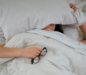 Why rest doesn’t feel restful. Image shows a tired woman in bed holding a pillow over her eyes 