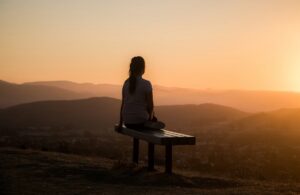 Image shows the back of a shadowed woman sat on a bench, looking at a sunset at the top of a hill 
