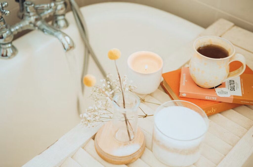 Image shows a bath with a tray over it containing a hot drink, a small case of flowers, books and a candle 